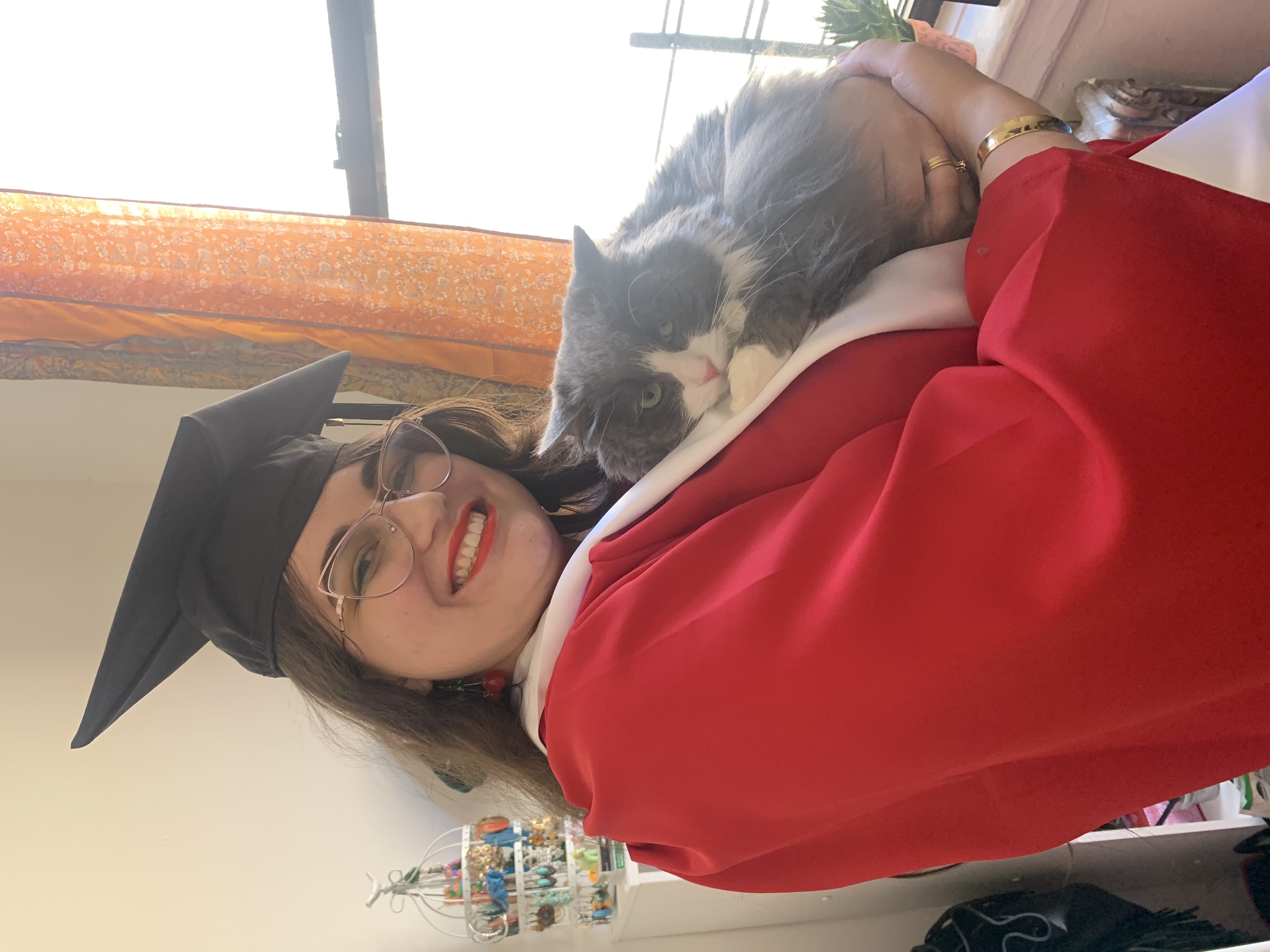 A white woman with long brown hair and cateye glasses wearing a red graduation robe and a black cap holding a long-haired gray-and-white cat with mint green eyes.