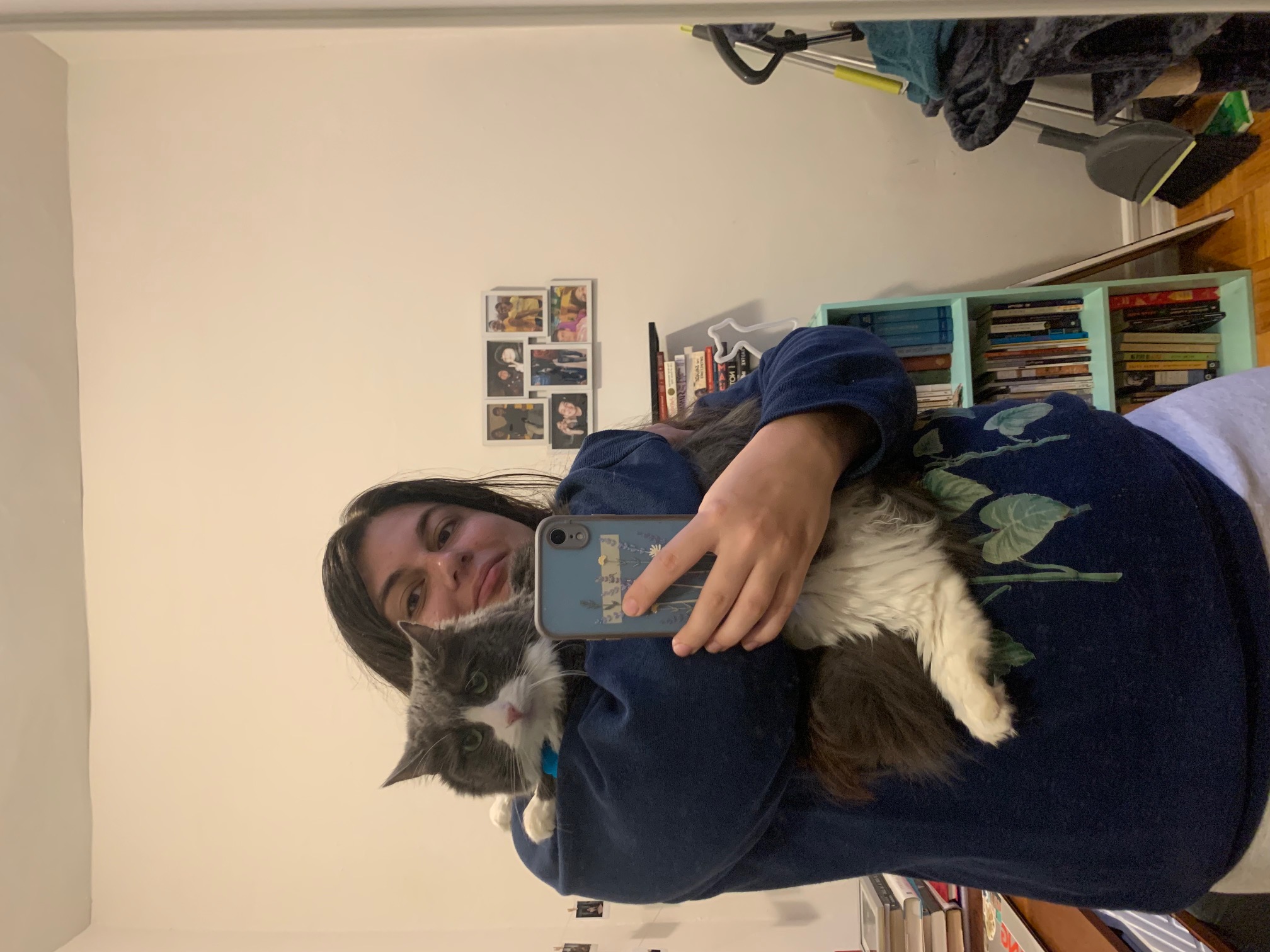 A mirror selfie of a white woman with brown hair wearing a sweatshirt and sweatpants and holding a long-haired gray-and-white cat with mint green eyes.